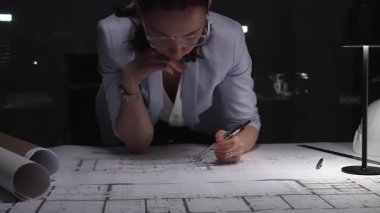 Profesyonel kadın mühendis inşaat projesini planlıyor, bina planlarını okuyor ve karanlık bir ofiste ayrıntıları düşünüyor..