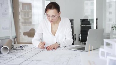 Kadın mimar, ayrıntılı bina planları üzerinde titizlikle çalışıyor. Masasına yayılmış. Mimari tasarımları parlak bir şekilde geliştirirken bir kalem ve cetvel kullanmak. Çağdaş ofis ortamı