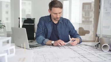 Profesyonel mimar modern bir masadaki detaylı yerleşim planlarını detaylı bir şekilde gözden geçirir. Laptop ve taslak araçları kullanarak kusursuz planlar yapar. Çağdaş bir ofiste teknolojiyle ev tasarımları yapılır.