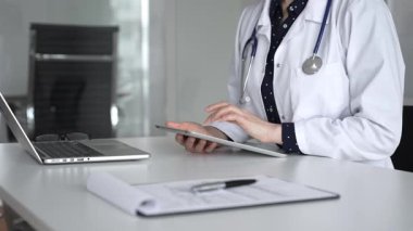 Modern dijital tablet kullanan beyaz önlüklü ve steteskoplu kadın doktor, klinik masasında hasta detayları üzerinde çalışıyor. Tıp, sağlık ve bilim konsepti