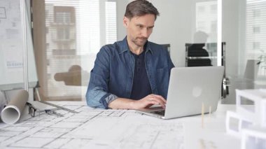 Profesyonel mimar modern bir masadaki detaylı yerleşim planlarını detaylı bir şekilde gözden geçirir. Laptop ve taslak araçları kullanarak kusursuz planlar yapar. Çağdaş bir ofiste teknolojiyle ev tasarımları yapılır.