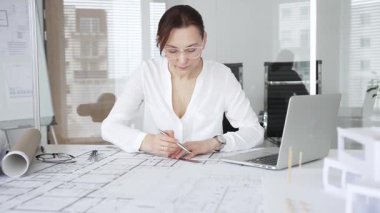 Kadın mimar, ayrıntılı bina planları üzerinde titizlikle çalışıyor. Masasına yayılmış. Mimari tasarımları parlak bir şekilde geliştirirken bir kalem ve cetvel kullanmak. Çağdaş ofis ortamı