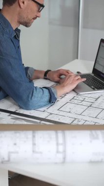 Mimar, yeni projeler için ayrıntılı planlama, inşaat ve tasarım mühendisliğini sembolize eden, kat planlarını gösteren bir cetvel ve dizüstü bilgisayar kullanarak beyaz bir masanın üzerine bina planları çiziyor.