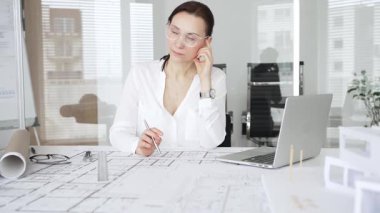 Masasında mimari planlar ve modeller üzerinde çalışan düşünceli bir kadın mimar elinde bir kalem tutuyor ve modern ofis ortamında proje detaylarını değerlendiriyor, kentsel gelişime odaklanıyor.