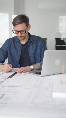 Erkek mimar masasına odaklanmış, dizüstü bilgisayardaki planları ve dijital planları gözden geçiriyor, modern ofis ortamında yenilikçi yerleşim ve kentsel tasarım çözümleri geliştiriyor.