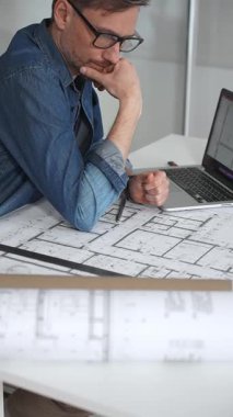 Mimar, yeni projeler için ayrıntılı planlama, inşaat ve tasarım mühendisliğini sembolize eden, kat planlarını gösteren bir cetvel ve dizüstü bilgisayar kullanarak beyaz bir masanın üzerine bina planları çiziyor.