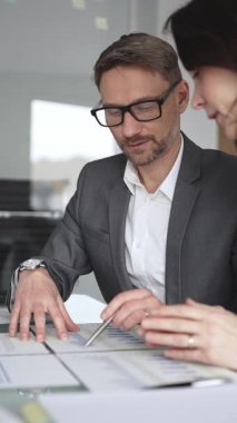 Kadın olarak masa başında işbirliği yapan iş adamları önemli bir sözleşme imzalarken, gözlüklü bir adam tablet üzerinde ayrıntılara dikkat çekiyor, takım çalışmasını ve karar verme mekanizmasını vurguluyor.