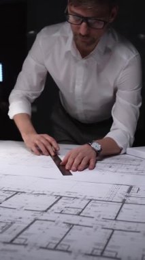Mimari tasarımlar üzerinde çalışan erkek mimar, büyük kağıt kağıtlara dikkatlice ölçüyor ve kat planlarını çiziyor. Gösterişsiz ışıklandırmada mimari ve iş kavramı