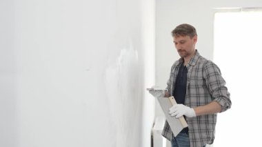 Koruyucu eldiven ve ekose gömlek giyen erkek inşaat işçisi, tadilat projesi sırasında profesyonel alçı levha bantlama bıçağı kullanarak duvarın yüzeyine sorunsuzca alçı yayıyor. Dikey görünüm.