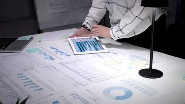 Mali raporlar ve çizelgelerle kaplı bir masada duran profesyonel iş adamı, bir akşam çalışması sırasında verileri analiz etmek ve kurumsal stratejiyi planlamak için dijital tabletle etkileşime geçiyor