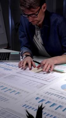 Mali tablolar, raporlar ve evrak işleriyle çevrili loş bir ofiste geç saatlere kadar çalışan genç bir profesyonel, hesaplama rakamları ve proje analizi ve planlamasını sonuçlandırmak için notlar alıyor.