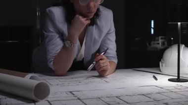 Profesyonel kadın mühendis inşaat projesini planlıyor, bina planlarını okuyor ve karanlık bir ofiste ayrıntıları düşünüyor..