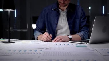 Geç saatlere kadar çalışan, ofis masasındaki dizüstü bilgisayardaki mali verileri ve iş raporlarını analiz eden, performans grafiklerini inceleyen ve planlama stratejisini lamba ışığında inceleyen, kurumsal büyümeyi yönlendiren bir adam.