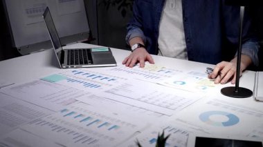 Mali tablolar, raporlar ve evrak işleriyle çevrili loş bir ofiste geç saatlere kadar çalışan genç bir profesyonel, hesaplama rakamları ve proje analizi ve planlamasını sonuçlandırmak için notlar alıyor.