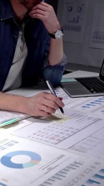 Gözlük takan, iş belgelerini ve dijital tableti inceleyen, strateji ve planlamayı düşünen, loş bir ofisteki lambayla aydınlatılmış bir masada fazla mesai yapan bir adam.