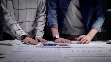 Bir masada iş birliği yapan iş adamları, kağıtlarda ve tablet ekranda gösterilen sayısız mali belge, grafik ve veriyi inceleyerek, ofis ortamında geç saatlere kadar çalışırlar.