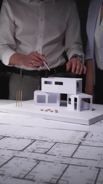 Mimarlar bir inşaat projesinde işbirliği yapıyor, mimari bir ev modeli ile çalışıyor ve bir masanın üzerinde bina planları hazırlıyor. Gösterişsiz ışıklandırmada mimari ve iş kavramı
