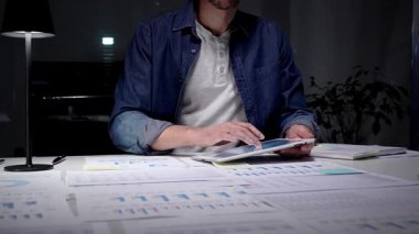 Gece geç saatlere kadar karanlık bir ofiste çalışan meşgul bir adam, stratejik planlama ve fazla mesai üretkenliği için dijital tablet kullanan finansal veriler, piyasa analizleri ve iş raporları üzerinde yoğunlaşıyor.