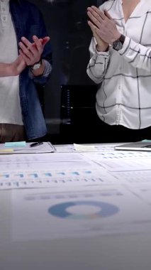 İş arkadaşları, mali belgeler, çizelgeler, tablet ve lambayla kaplı ofis masasını alkışlayarak, gece yarısı yapılan analizlerden sonra anlaşmaya işaret ediyor ve başarılı sonuçları kutluyorlar.