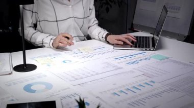 Geceleri titizlikle çalışan, sayısız basılı çizelge, grafik ve dizüstü bilgisayarla çevrili, henüz düzenlenmemiş bir masanın mali verilerini ve iş raporlarını titizlikle analiz eden bir kişi.