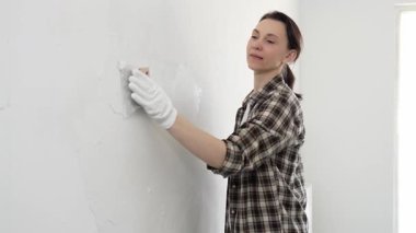 Koruyucu eldiven giyen ve kareli gömlekli bir kadın beyaz duvara macun yapıştırıyor, ev tadilatı sırasında spatulayla bileşik yayıyor, pratik ev geliştirme becerileri sergiliyor..