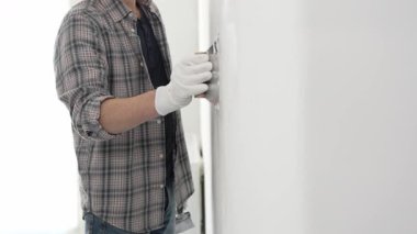 Eldiven ve ekose gömlek giyen tanınmamış inşaat işçisi, bir ev yenileme veya inşaat projesi sırasında beyaz bir duvara sıva uygulamak için macun bıçağı kullanıyor..