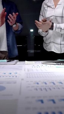 İş arkadaşları, mali belgeler, çizelgeler, tablet ve lambayla kaplı ofis masasını alkışlayarak, gece yarısı yapılan analizlerden sonra anlaşmaya işaret ediyor ve başarılı sonuçları kutluyorlar.