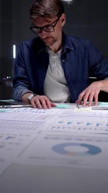 Gece geç saatlere kadar karanlık bir ofiste çalışan meşgul bir adam, stratejik planlama ve fazla mesai üretkenliği için dijital tablet kullanan finansal veriler, piyasa analizleri ve iş raporları üzerinde yoğunlaşıyor.