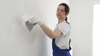 Profesyonel inşaat işçisi kadın, beyaz bir duvarı onarmak için macun bıçağı kullanıyor, bir kat macun sürüyor ve temiz bir iş için beyaz eldiven takıyor.