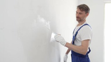 Eldiven ve mavi tulum giyen orta yaşlı inşaat işçisi, beyaz bir duvara sıva uygulamak için macun bıçağı kullanıyor. Ev yenileme ve iyileştirme yapıyor..