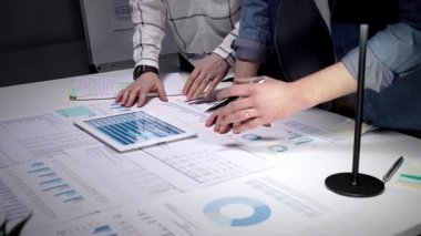 İş dünyasından profesyonellerin elleri dijital tabletle etkileşime girerek çizelgeleri ve çeşitli mali belgeler, evraklar ve raporlarla kaplı bir masayı bir takım toplantısında analiz ediyorlar.