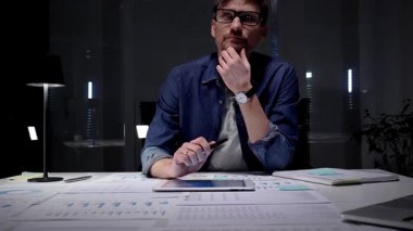 Gözlük takan, iş belgelerini ve dijital tableti inceleyen, strateji ve planlamayı düşünen, loş bir ofisteki lambayla aydınlatılmış bir masada fazla mesai yapan bir adam.
