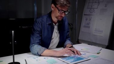 Geceleyin işe dalmış, mali belgeleri ve istatistikleri inceliyor, veri işlemek için bir tablet kullanıyor, teslim tarihlerine ve uzun saatlere olan bağlılığını vurguluyor.