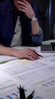 Gözlük takan, iş belgelerini ve dijital tableti inceleyen, strateji ve planlamayı düşünen, loş bir ofisteki lambayla aydınlatılmış bir masada fazla mesai yapan bir adam.