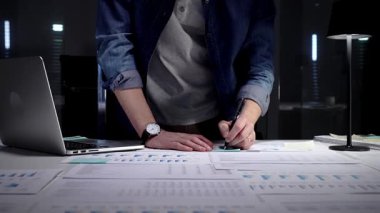 Geç saatlere kadar çalışan girişimci, kağıt, rapor ve dizüstü bilgisayarlardaki iş istatistikleri ve mali verileri analiz ederek, geceleri loş işyerinde kurumsal strateji ve gelecekteki büyümeyi yönetmektedir.