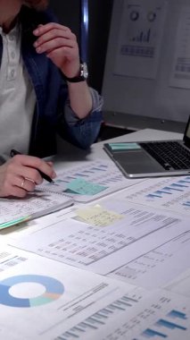 Mali tablolar, raporlar ve evrak işleriyle çevrili loş bir ofiste geç saatlere kadar çalışan genç bir profesyonel, hesaplama rakamları ve proje analizi ve planlamasını sonuçlandırmak için notlar alıyor.