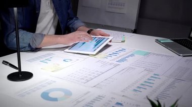 Dijital tablet kullanarak finansal verileri analiz eden profesyonel bir adam. İş raporlarını yönetiyorum. Çizelgeler. Ve yoğun bir ofis masasında, ışık altında istatistikler. Doğru stratejik planlamanın sağlanması