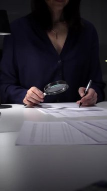 Vergi formlarını ve mali beyannameleri inceleyen kadın, hesap makinesi ve büyüteç kullanarak rakamları ve denetim kayıtlarını kontrol ediyor. İş dünyasında denetim ve vergi teması