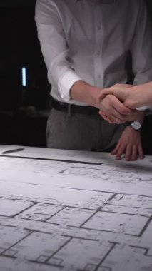 Profesyoneller planların üzerinden el sıkışıyor, bir inşaat projesi için iş anlaşmasını sonuçlandırıyor, mimari planlama ortaklığı. Gösterişsiz ışıklandırmada mimari ve iş kavramı