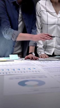 Bir masada iş birliği yapan iş adamları, kağıtlarda ve tablet ekranda gösterilen sayısız mali belge, grafik ve veriyi inceleyerek, ofis ortamında geç saatlere kadar çalışırlar.