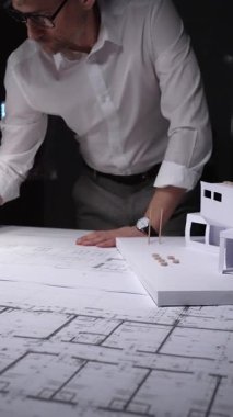Profesyonel mimar veya mühendis bina tasarımını geliştiriyor, kat planları ve bir masanın üzerinde ölçeklendirilmiş mimari modelle çalışıyor. Gösterişsiz ışıklandırmada mimari ve iş kavramı