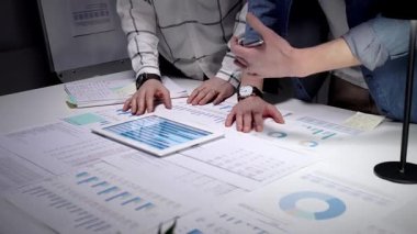 İş dünyasından profesyonellerin elleri dijital tabletle etkileşime girerek çizelgeleri ve çeşitli mali belgeler, evraklar ve raporlarla kaplı bir masayı bir takım toplantısında analiz ediyorlar.