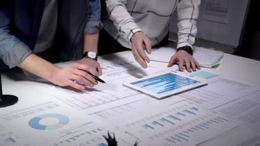 İş dünyasından profesyonellerin elleri dijital tabletle etkileşime girerek çizelgeleri ve çeşitli mali belgeler, evraklar ve raporlarla kaplı bir masayı bir takım toplantısında analiz ediyorlar.