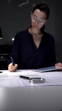 Tablet verilerini analiz eden ve loş bir ofis lambasının altındaki masada evrakları imzalayan bir kadın, gece geç saatlerde raporlara ve sözleşmelere odaklandı. İş dünyasında denetim ve vergi teması
