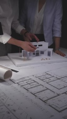 Modern bir ev tasarımında işbirliği yapan mimarlar, karanlık bir masaüstünde ölçekli bir model ve mühendislik planlarıyla çalışıyorlar. Gösterişsiz ışıklandırmada mimari ve iş kavramı