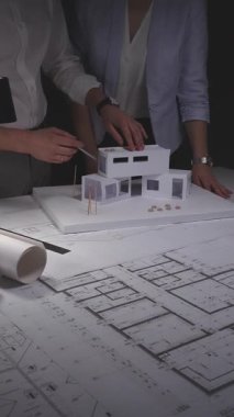 Modern bir ev tasarımında işbirliği yapan mimarlar, karanlık bir masaüstünde ölçekli bir model ve mühendislik planlarıyla çalışıyorlar. Gösterişsiz ışıklandırmada mimari ve iş kavramı
