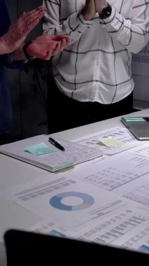 İş arkadaşları, mali belgeler, çizelgeler, tablet ve lambayla kaplı ofis masasını alkışlayarak, gece yarısı yapılan analizlerden sonra anlaşmaya işaret ediyor ve başarılı sonuçları kutluyorlar.