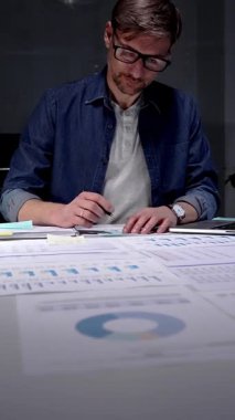 Gece geç saatlere kadar karanlık bir ofiste çalışan meşgul bir adam, stratejik planlama ve fazla mesai üretkenliği için dijital tablet kullanan finansal veriler, piyasa analizleri ve iş raporları üzerinde yoğunlaşıyor.