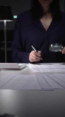 Vergi formlarını ve mali beyannameleri inceleyen kadın, hesap makinesi ve büyüteç kullanarak rakamları ve denetim kayıtlarını kontrol ediyor. İş dünyasında denetim ve vergi teması