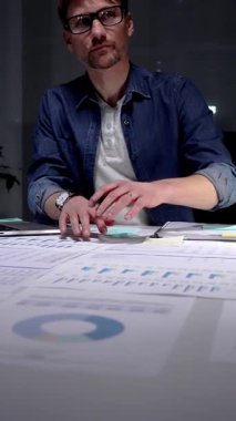 Gece geç saatlere kadar karanlık bir ofiste çalışan meşgul bir adam, stratejik planlama ve fazla mesai üretkenliği için dijital tablet kullanan finansal veriler, piyasa analizleri ve iş raporları üzerinde yoğunlaşıyor.